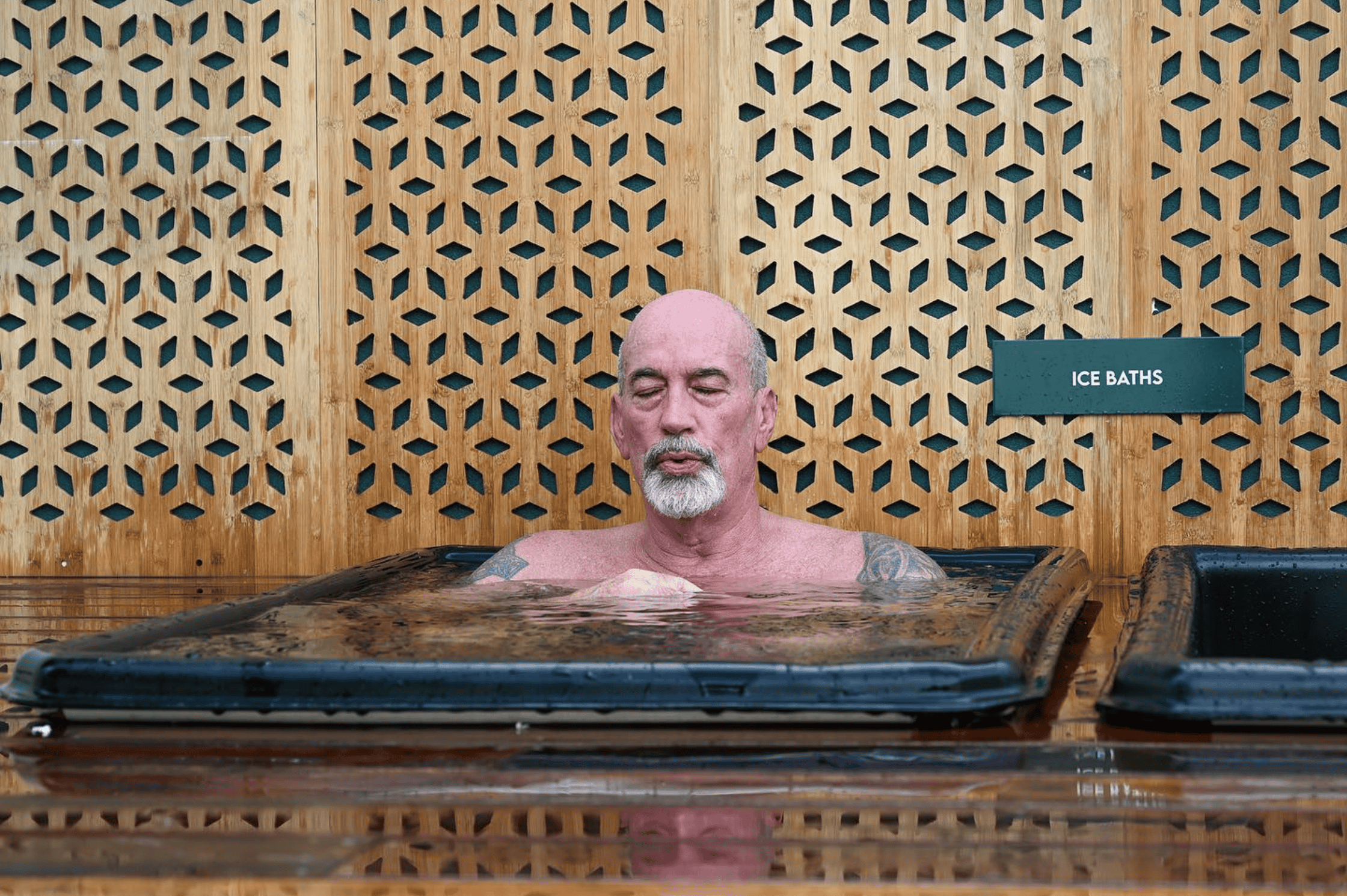 Man sitting in an ice bath, eyes closed, wooden wall in the background.